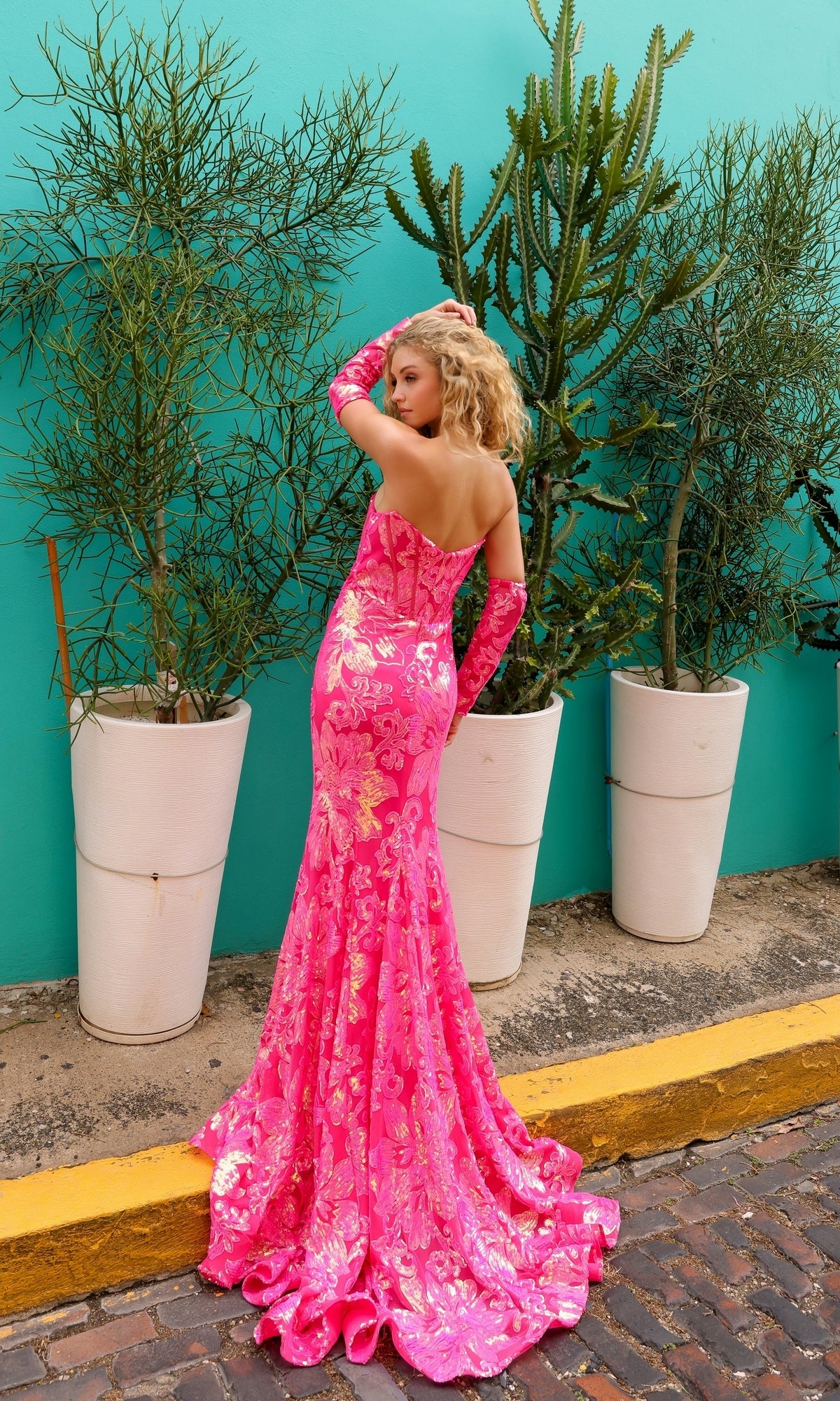 A woman poses outdoors in front of potted plants and a turquoise wall, wearing the Strapless Sequin-Print Prom Dress R1268.